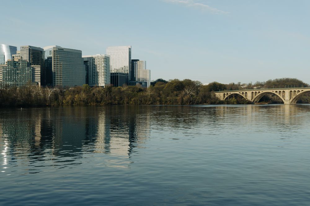 A scenic view of skyline buildings and Key Bridge reflecting on the Potomac River.