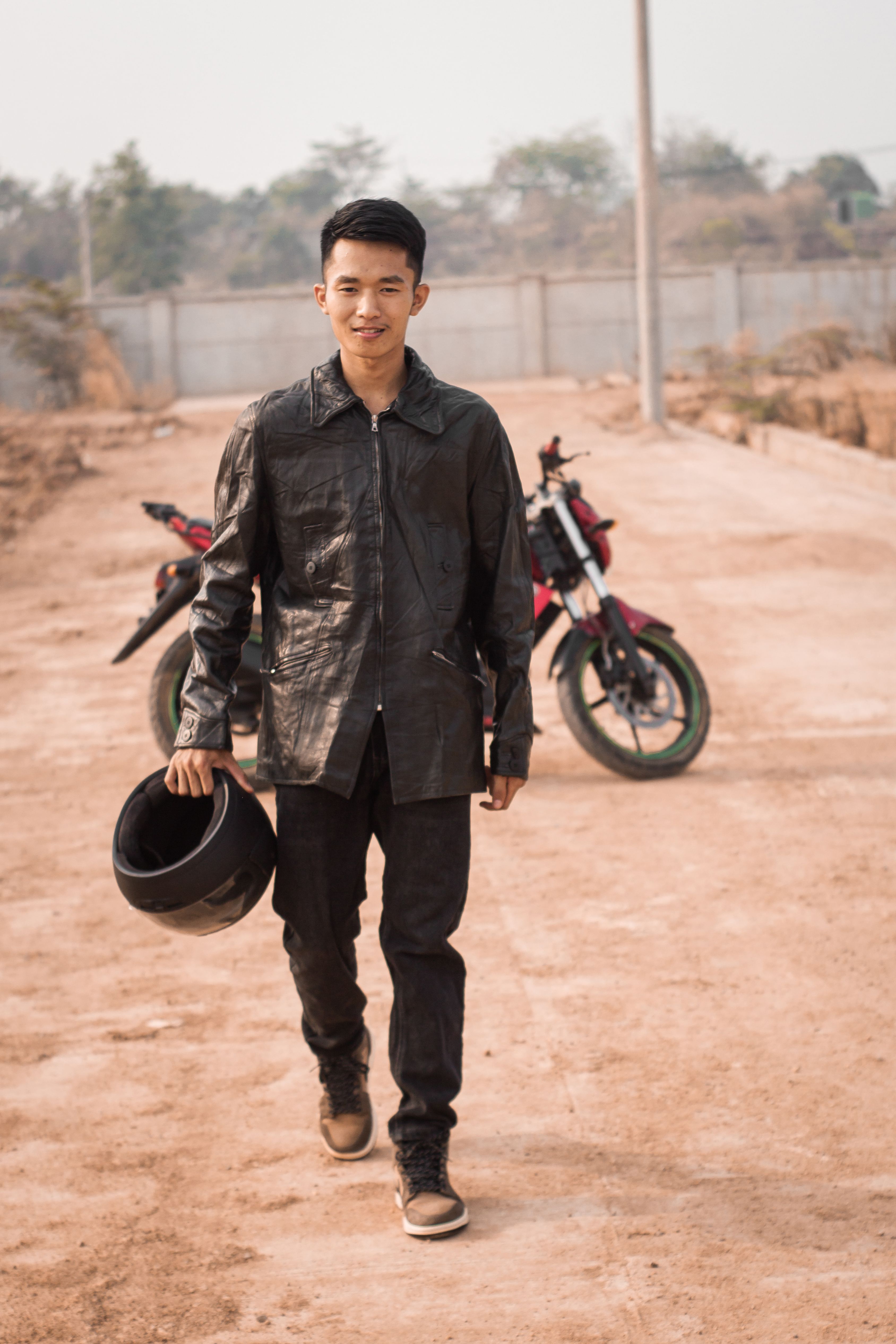 Young man in leather jacket walks confidently towards motorcycle on a dusty outdoor path.