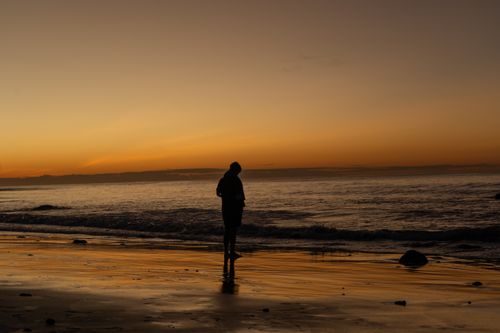 Free A silhouette walking along the beach at sunset, reflecting on water. Stock Photo