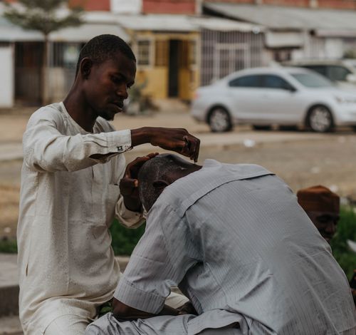 Free A street barber performs a haircut in an urban environment with a car in the background. Stock Photo