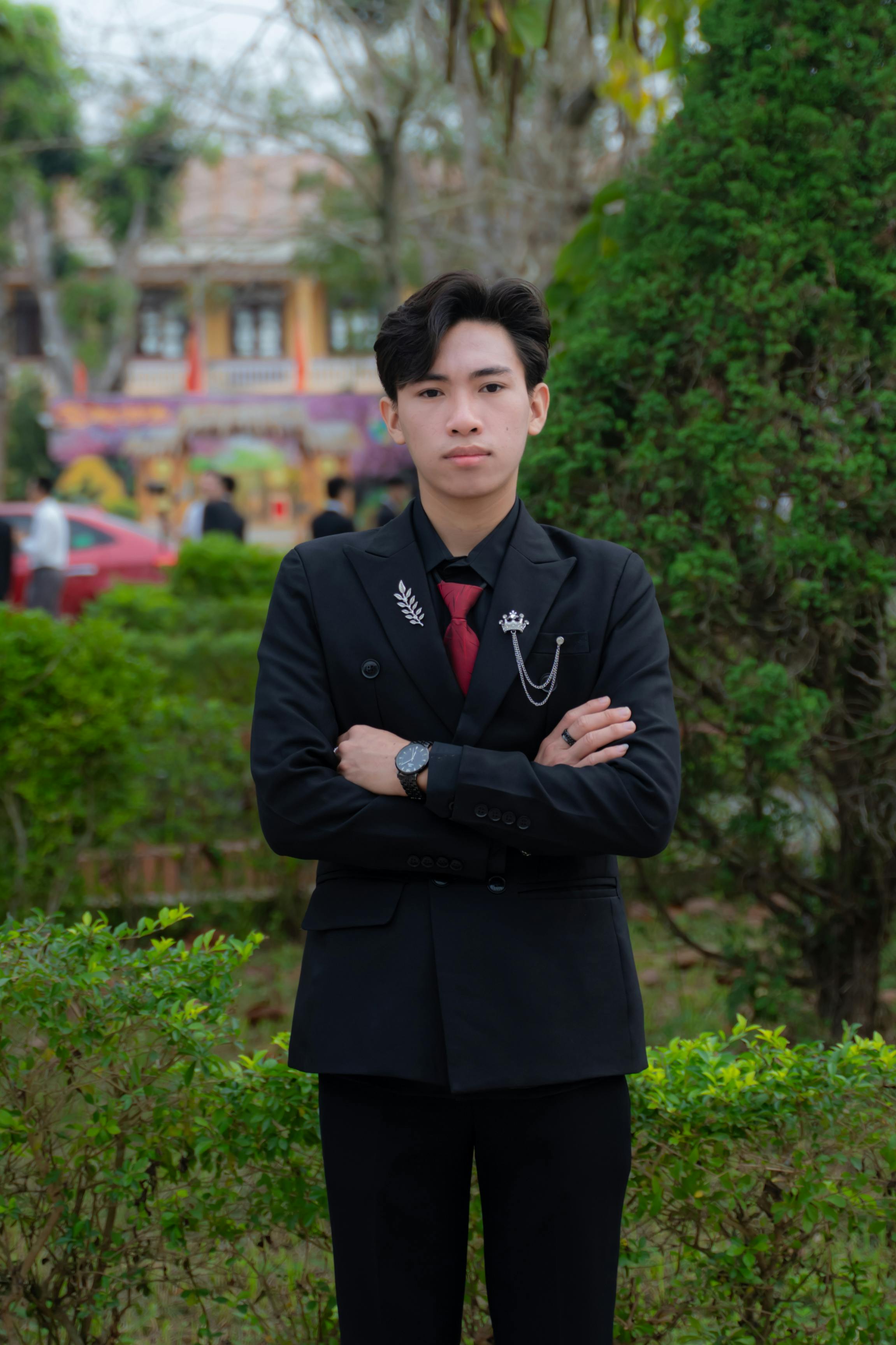 Young man stands confidently in a park wearing formal black suit with red tie.