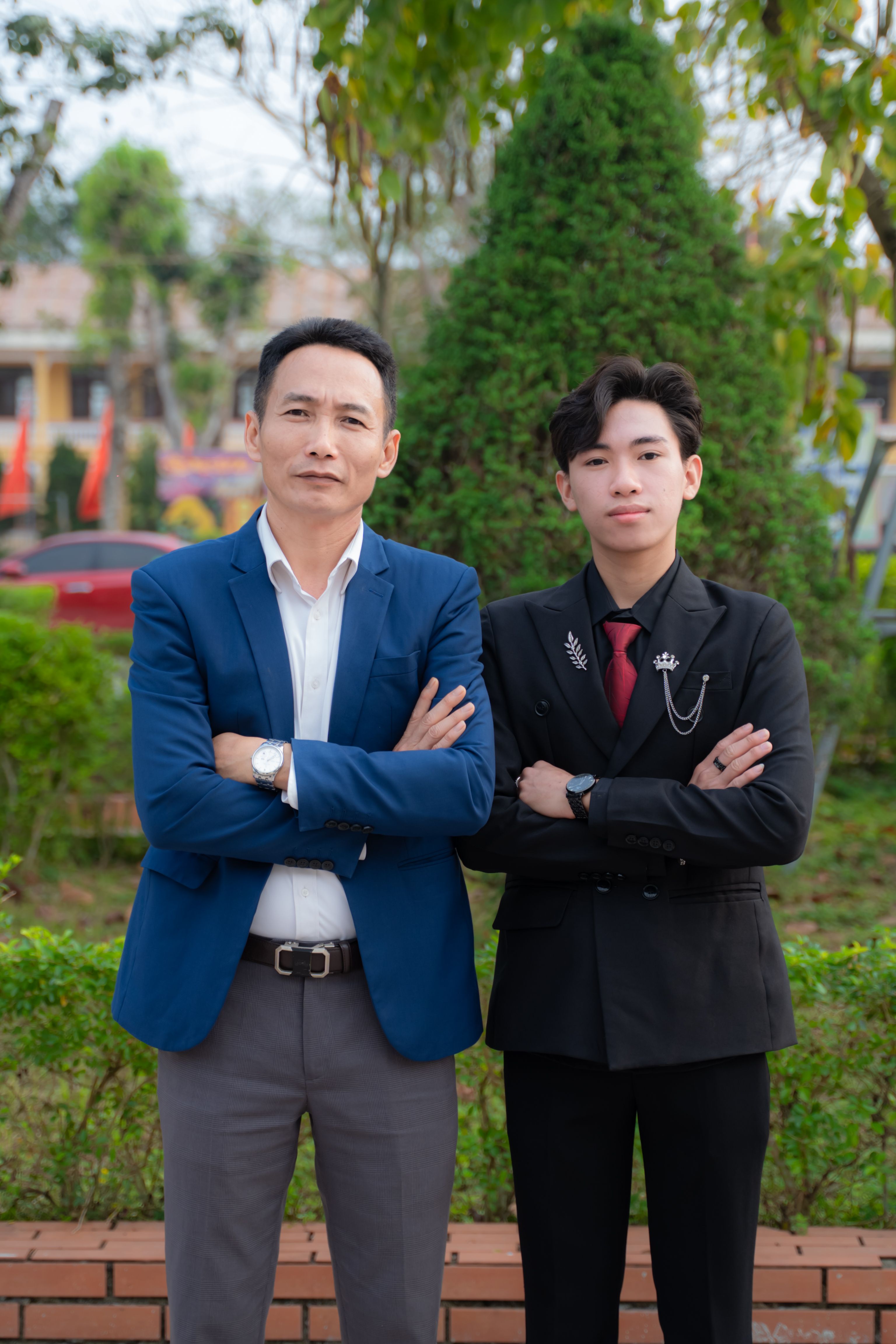 Father and teenage son in suits standing confidently outdoors with arms crossed.