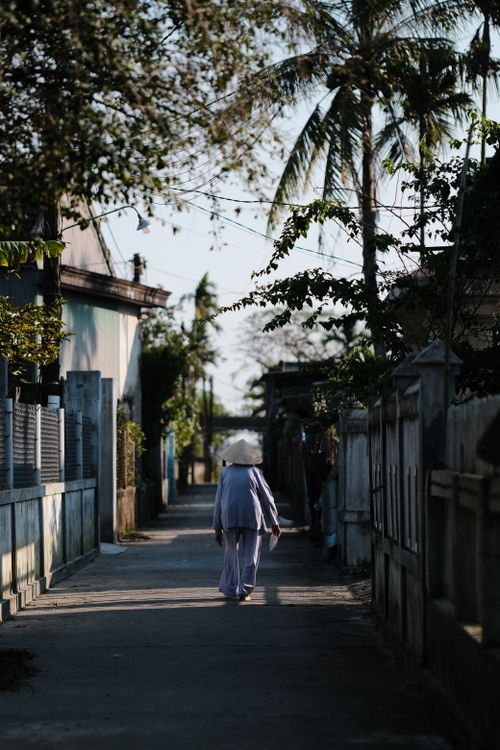 Free A person strolls through a tranquil village street lined with trees and houses, under a clear sky. Stock Photo