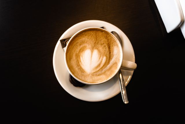 Overhead view of a flat white in a white cup and saucer on a dark table, with leaf-shaped foam art. Photo by Zulfugar Karimov on Pexels.