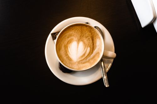 Free Overhead shot of a latte in a white cup with heart-shaped foam art on a dark table. Stock Photo