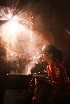 A woman in traditional attire cooking with a steaming pot, beautifully lit by soft window light.