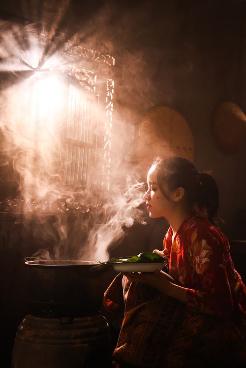 Free A woman in traditional attire cooking with a steaming pot, beautifully lit by soft window light. Stock Photo