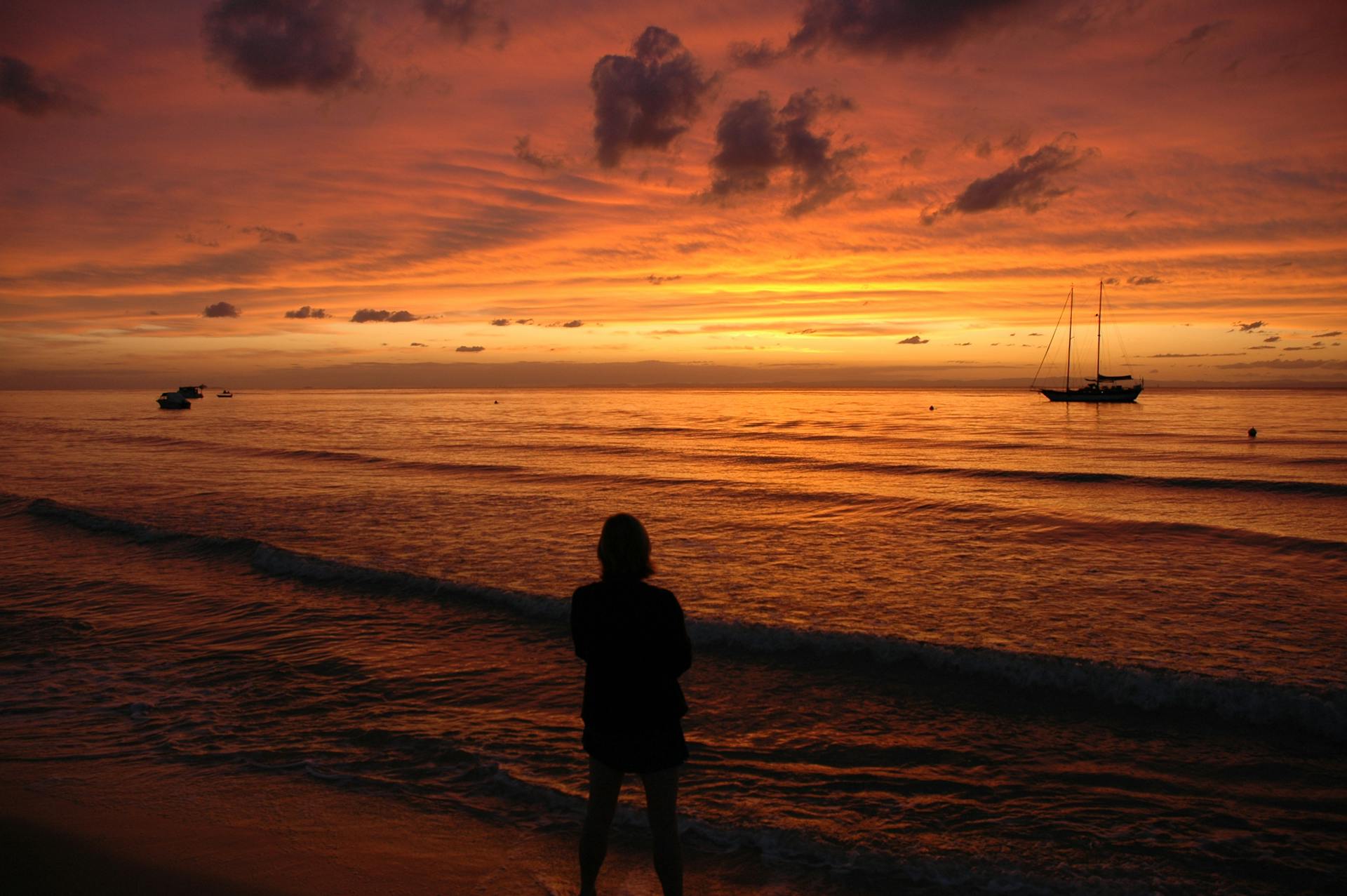Silhouette of a person watching sunset at Moreton Island beach with boats in view, creating a serene atmosphere.