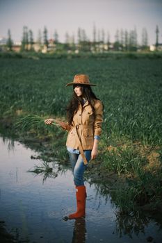 Young woman in rural field wearing coat, hat, and red boots, holding plants.