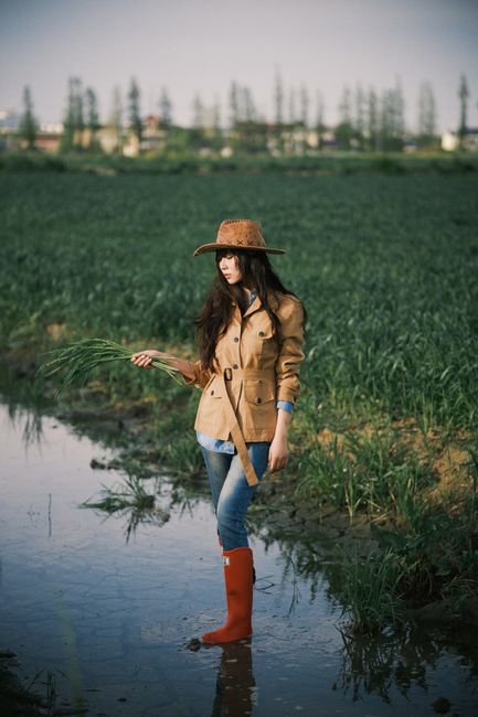 Young woman in rural field wearing coat, hat, and red boots, holding plants.