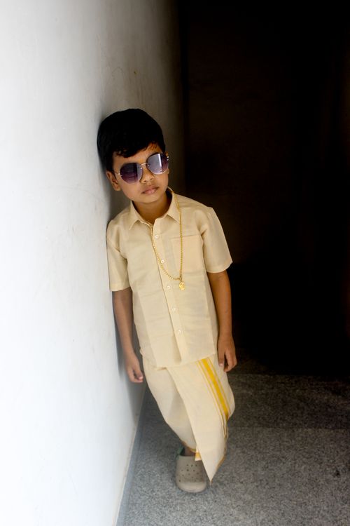 Free A young boy in traditional attire leans against a wall, wearing sunglasses and jewelry. Stock Photo