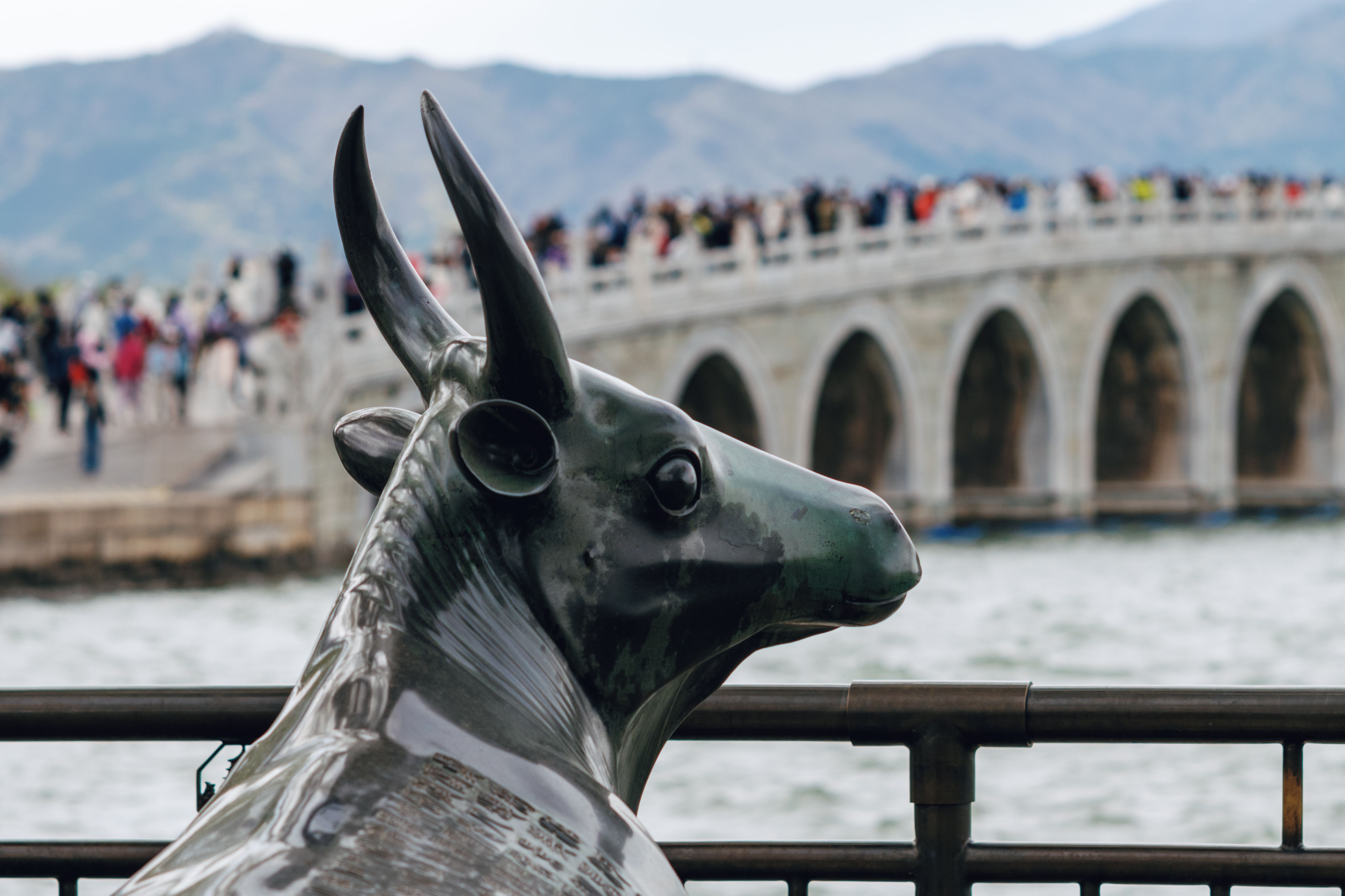 Bronze ox statue in front of the 17-Arch Bridge at Summer Palace, Beijing, featuring scenic view and crowd.