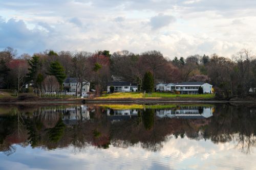 Free Charming lakeside homes reflect on a tranquil lake, surrounded by early spring foliage and a calm sky. Stock Photo