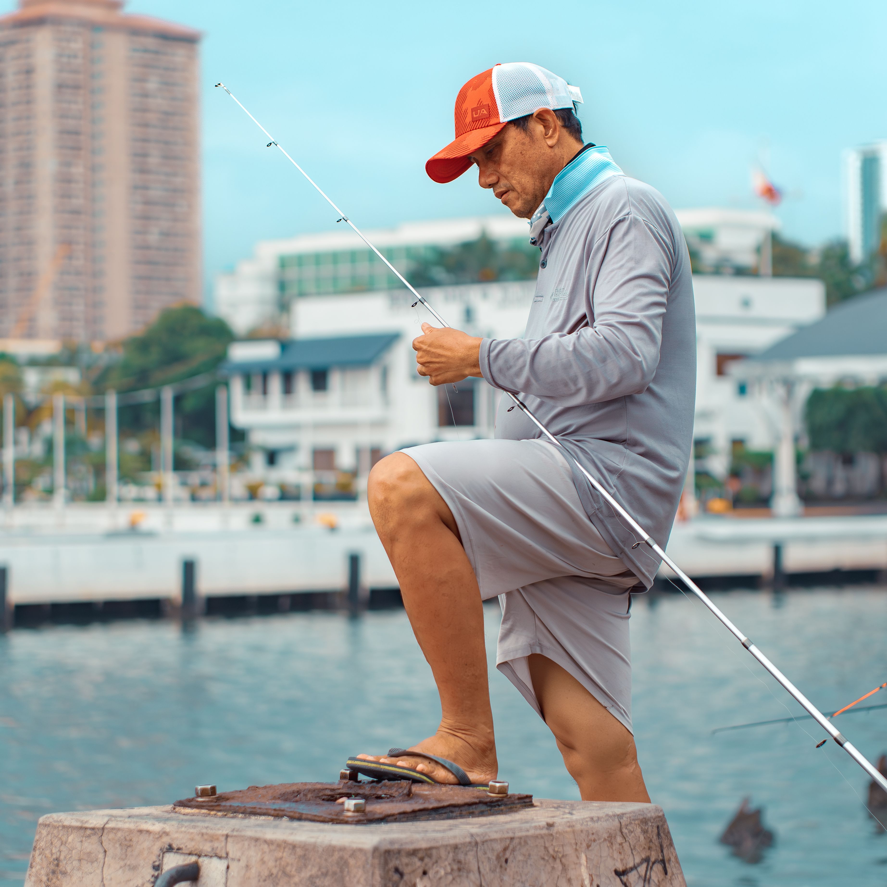 A man is fishing by a city waterfront, dressed in casual attire, against an urban backdrop.