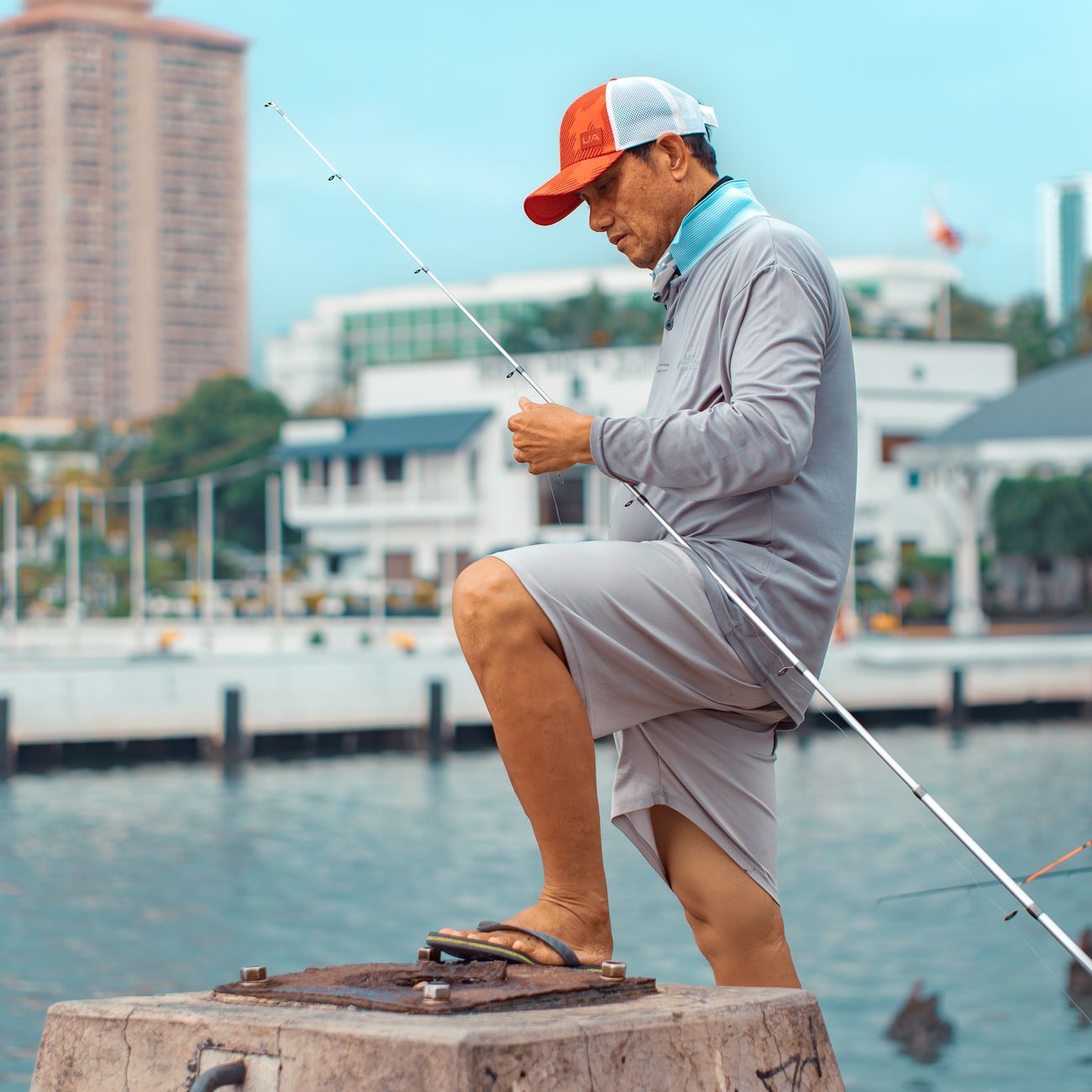 A man is fishing by a city waterfront, dressed in casual attire, against an urban backdrop.