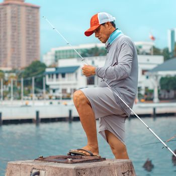 A man is fishing by a city waterfront, dressed in casual attire, against an urban backdrop.