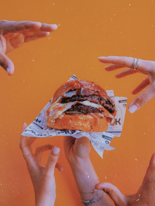 Free A close-up of hands reaching out to a mouth-watering burger against a vibrant orange background. Stock Photo