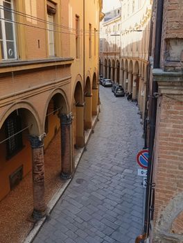 A sunny day on a quaint, historic street under classic Italian porticoes in Bologna.