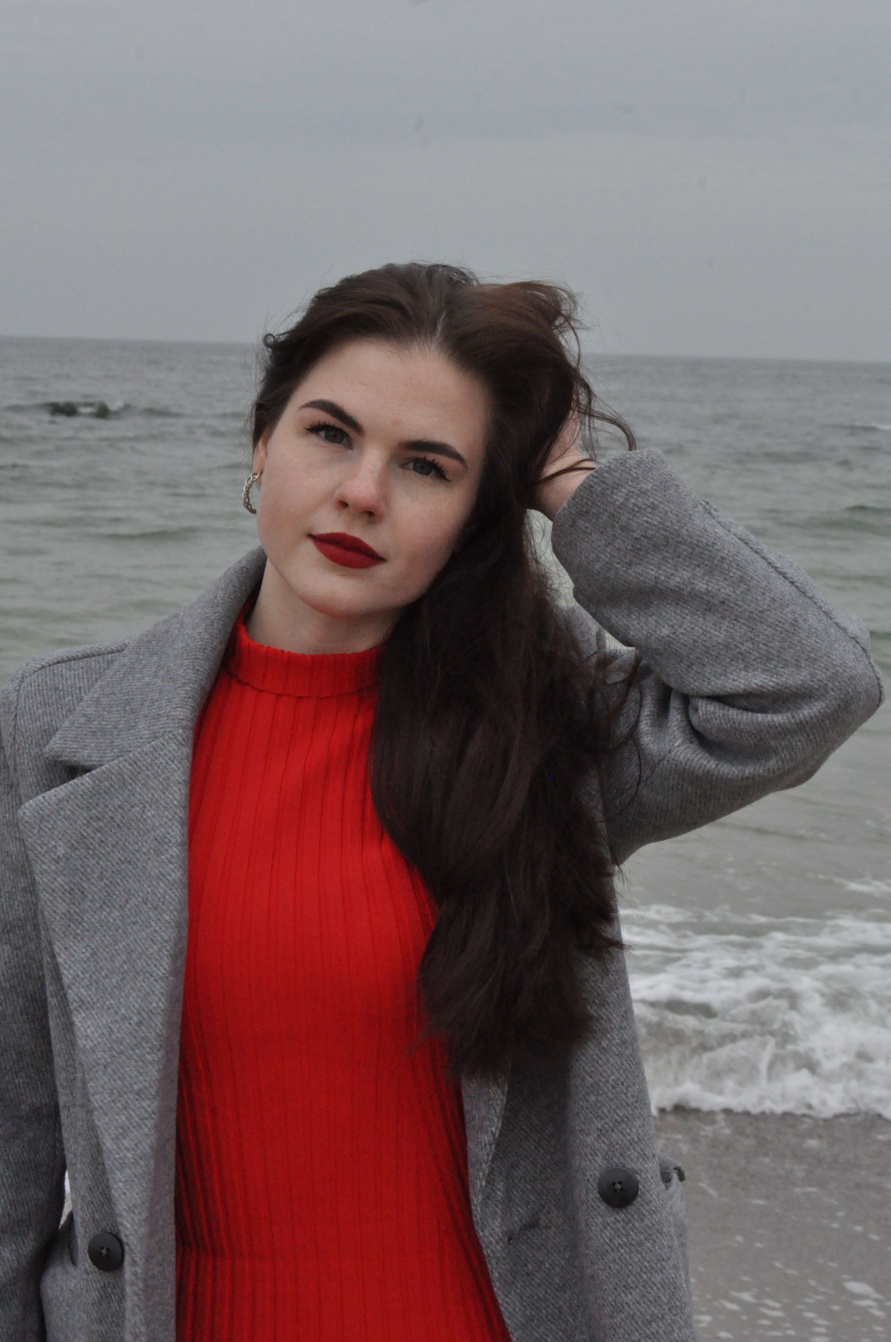Elegantly dressed woman in red and grey coat posing by the ocean.