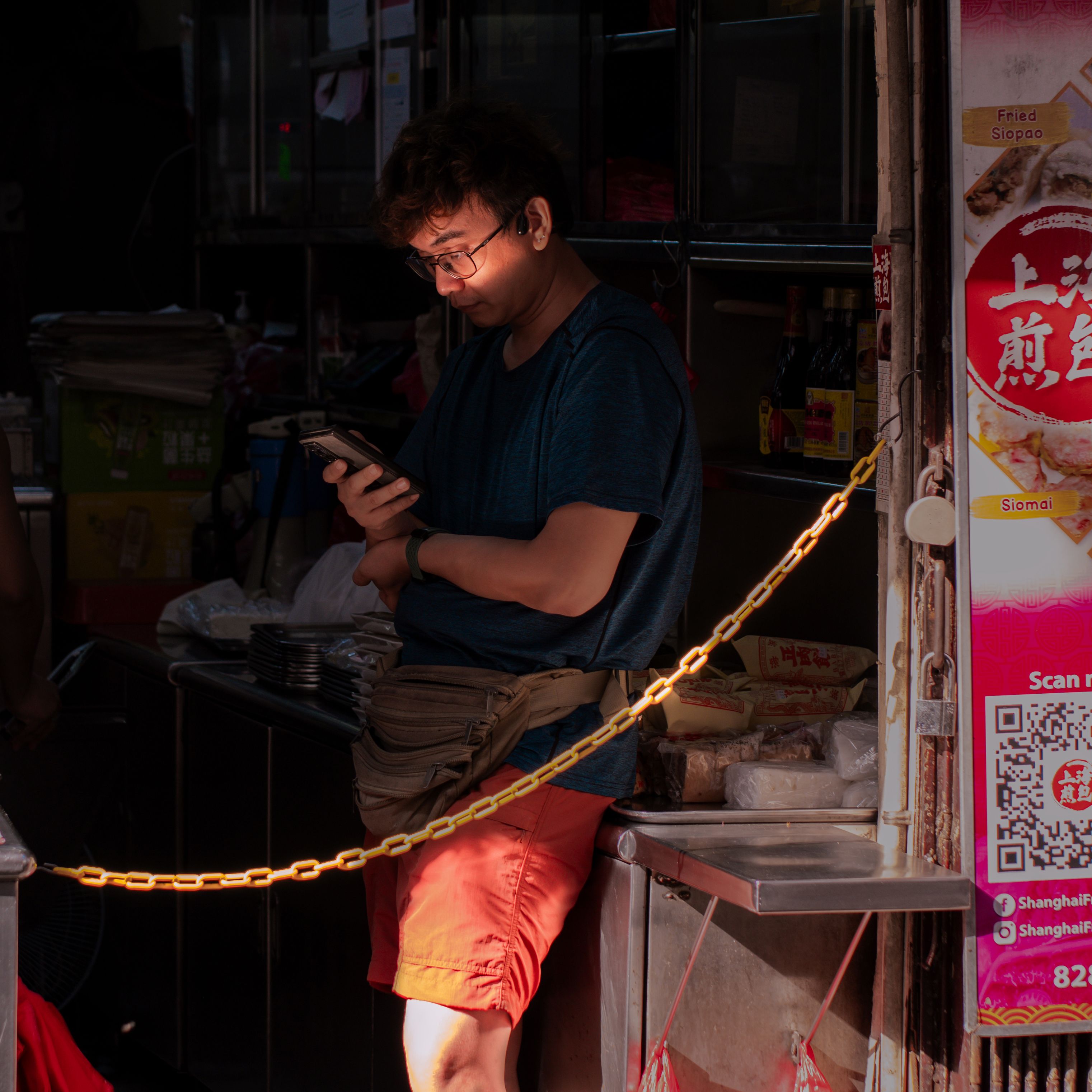 A man stands in a market stall using his phone, illuminated by sunlight.
