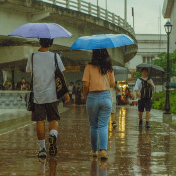 Street scene of people walking with umbrellas in the rain under a bridge.