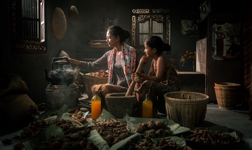 Free A mother and daughter engaged in traditional cooking inside, surrounded by ginger and baskets. Stock Photo