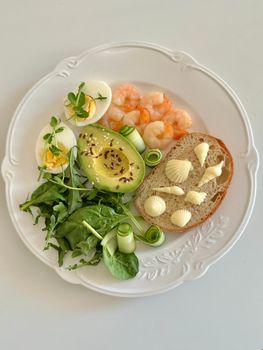 A gourmet plate featuring shrimp, avocado, eggs, greens, and bread arranged artfully on a white plate.