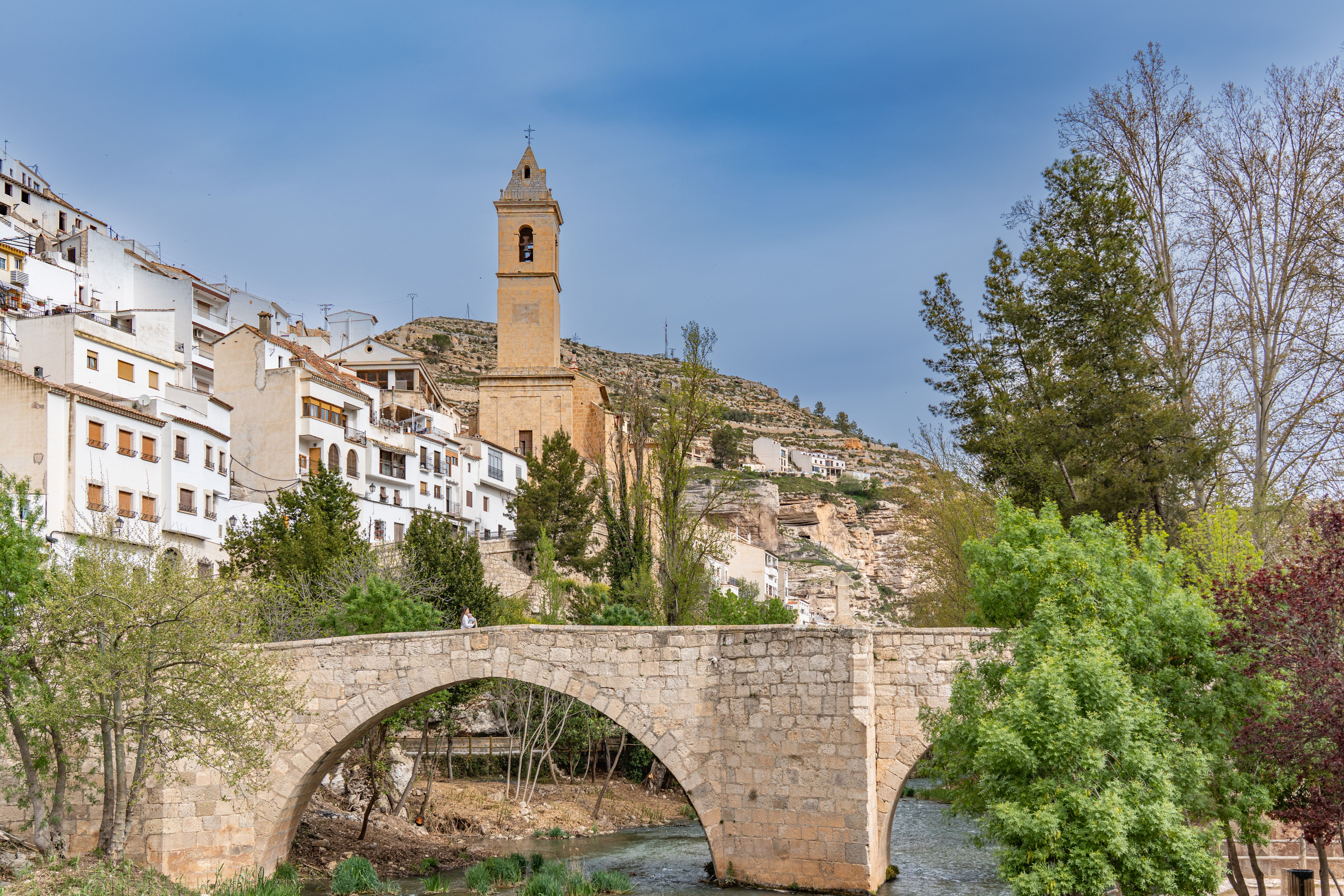 Scenic view of Alcalá del Júcar with its iconic stone bridge and church tower amidst lush greenery.