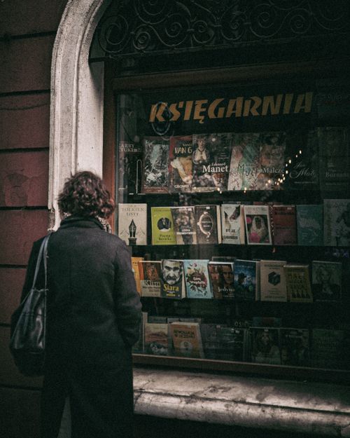 Free A woman in a dark coat browsing books displayed in a rustic bookstore window. Vintage atmosphere. Stock Photo