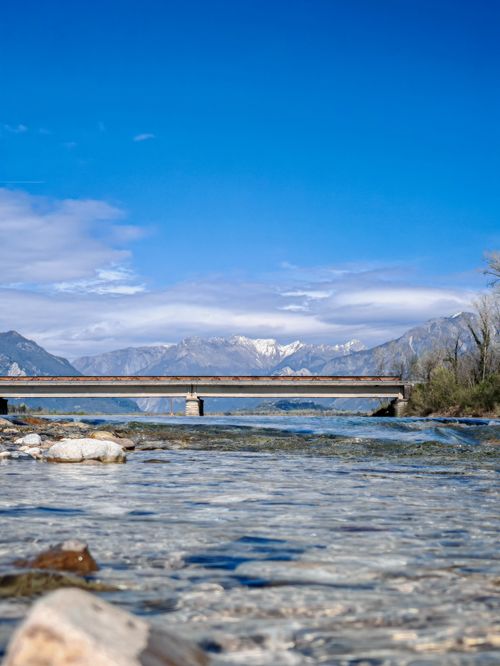 Free A picturesque view of a bridge over a tranquil river with snow-capped mountains in the background. Stock Photo