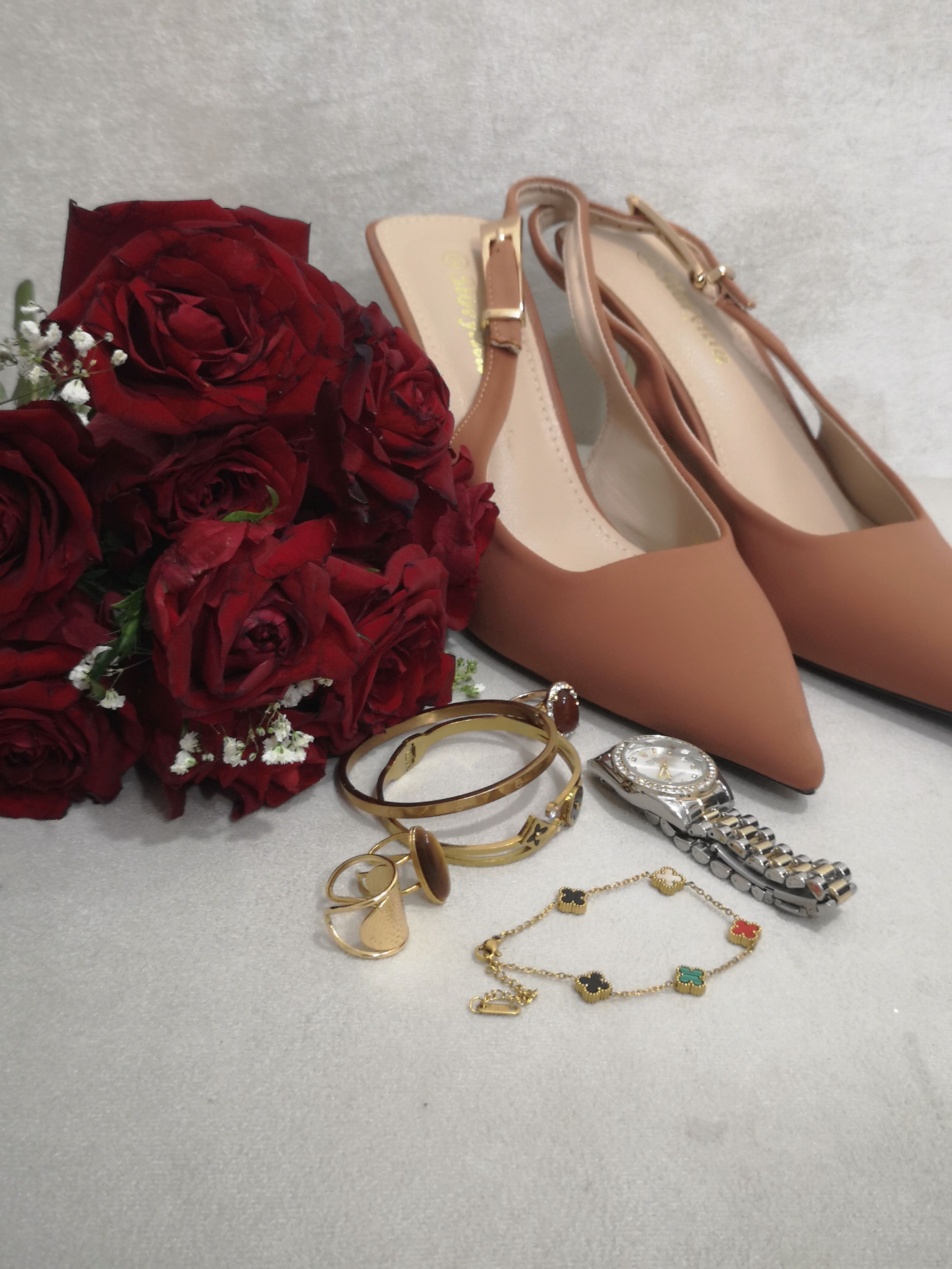 Arranjo sofisticado de sapatos de salto alto, joias e um buquê de rosas vermelhas sobre um fundo neutro.