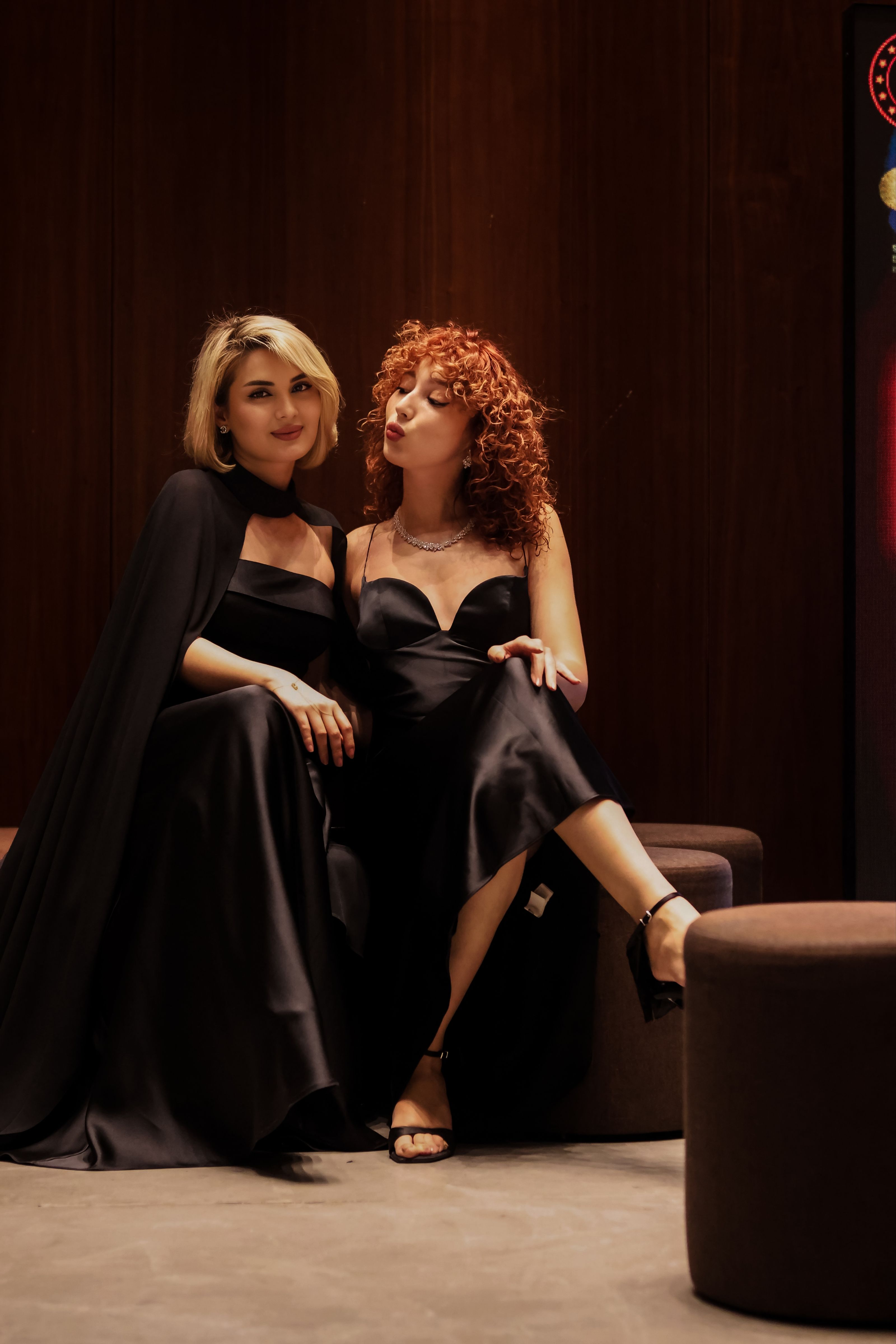 Duas mulheres em elegantes vestidos de noite pretos, sentadas em um ambiente interno, exalam elegância e sofisticação.