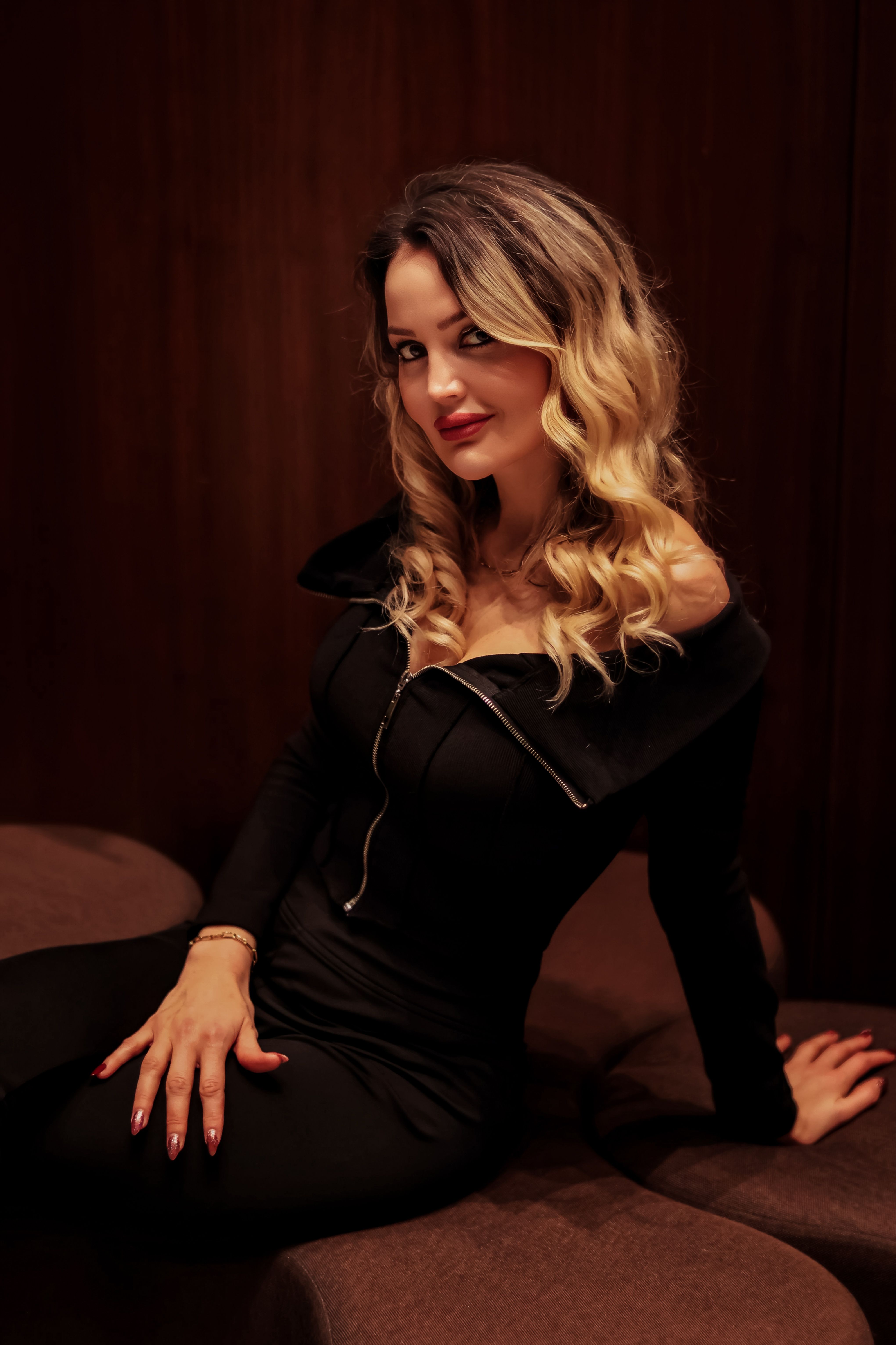 Stylish woman with blonde curls seated gracefully in a black dress, radiating elegance.