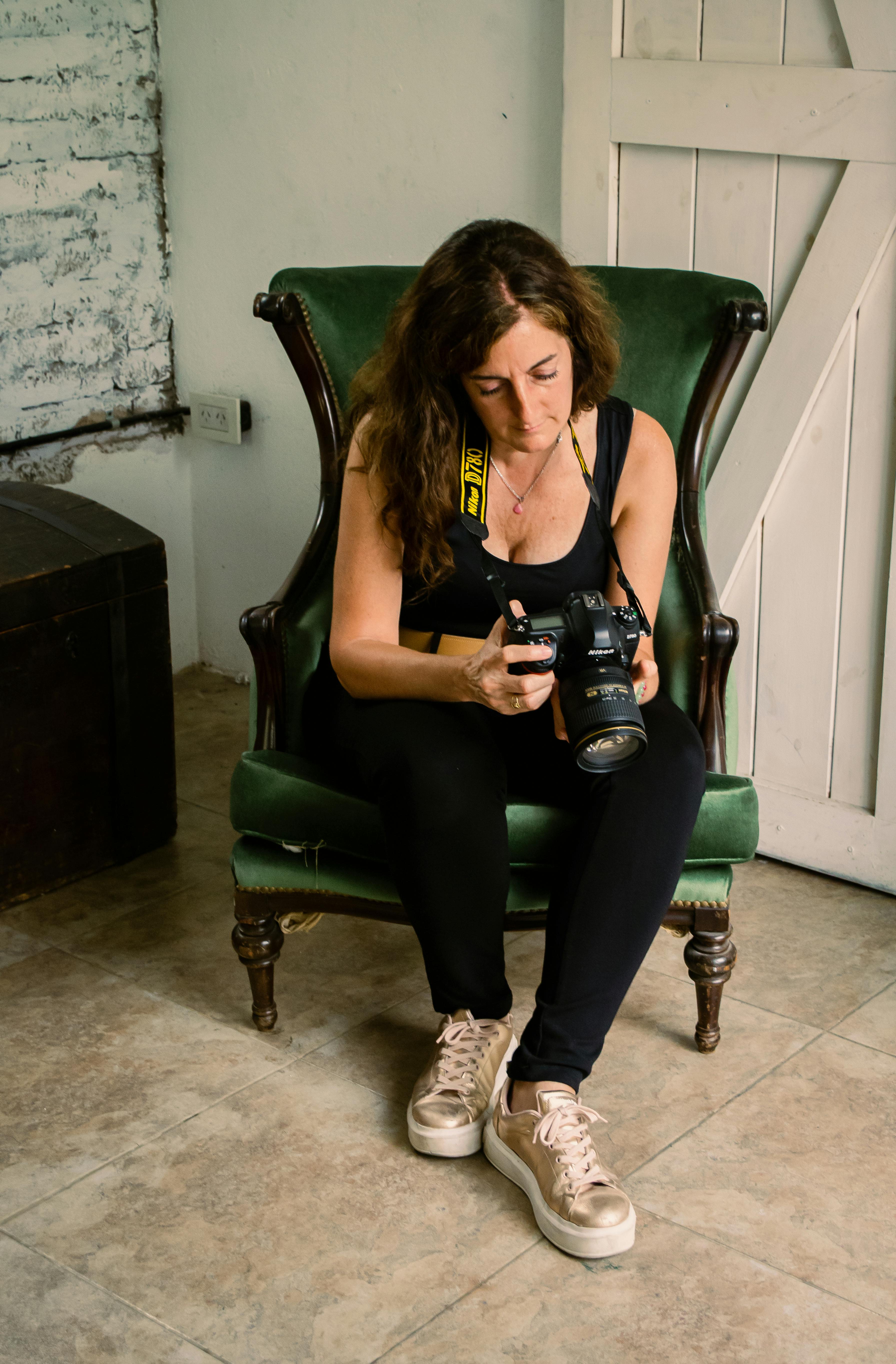 Woman sitting on green chair indoors, examining her camera. Rustic and creative atmosphere with vintage accents.