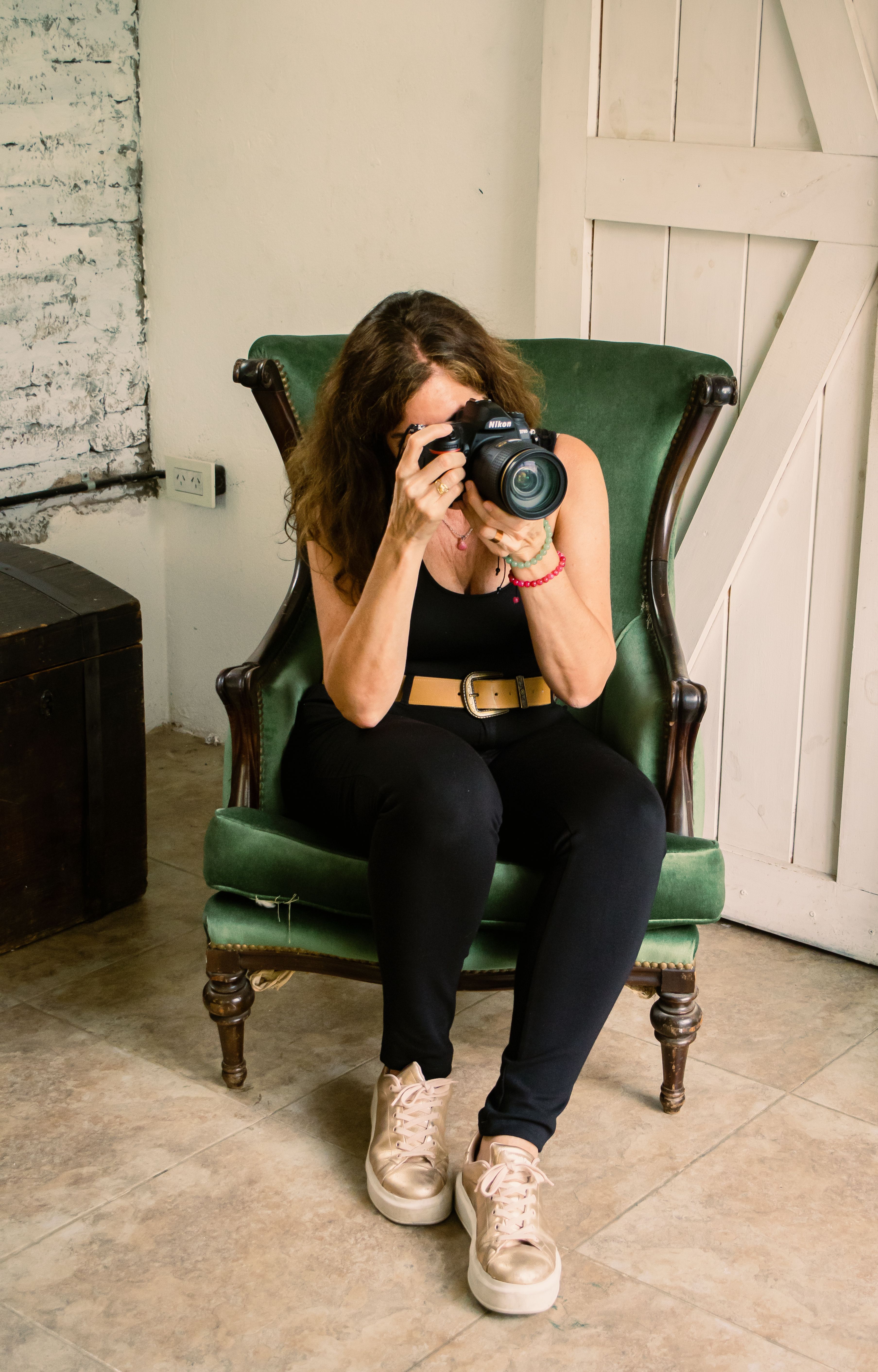 A woman sits in a green armchair indoors, taking a photo with a camera.