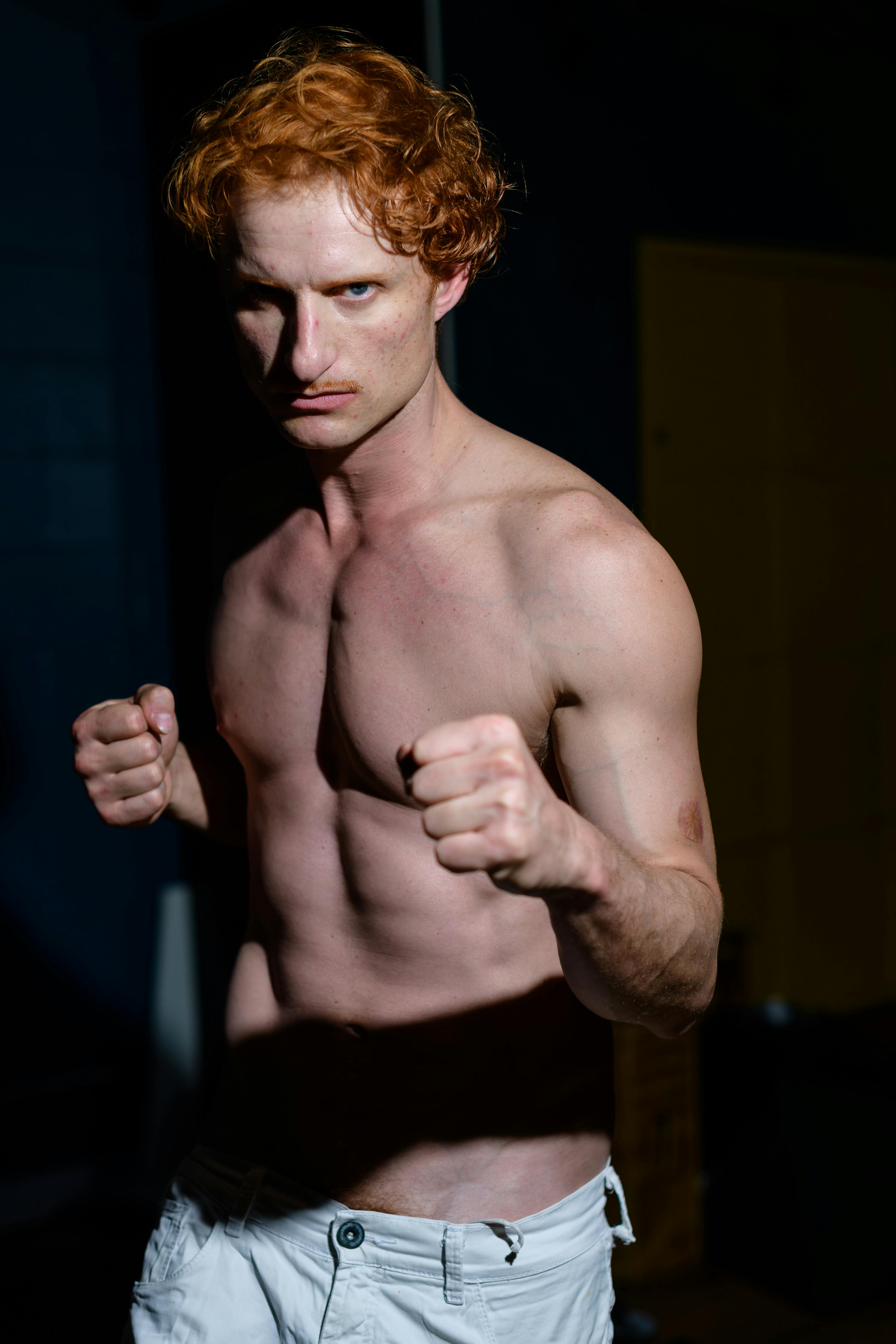 Shirtless man with red hair poses in a boxing stance indoors with dramatic lighting.