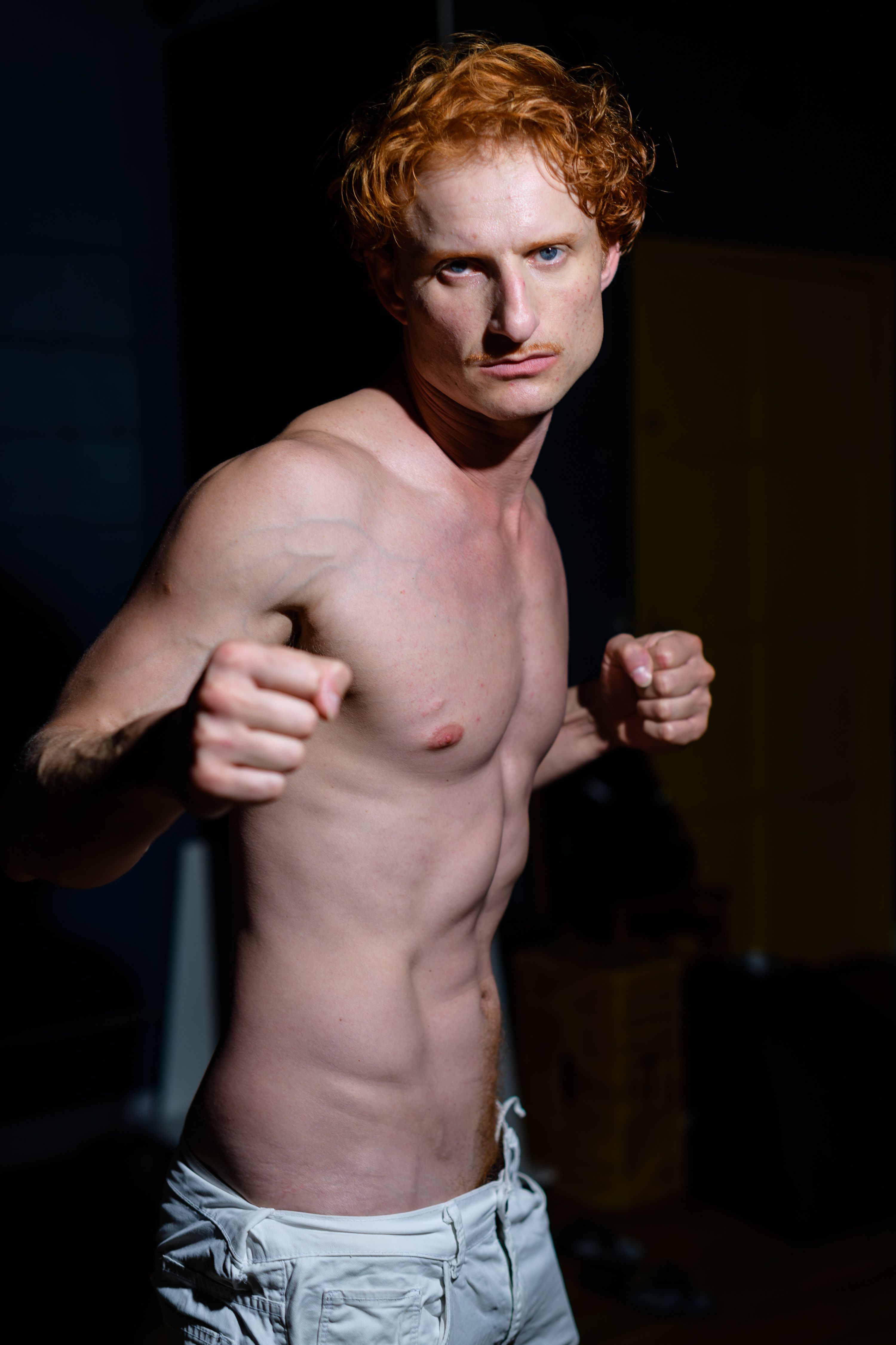 Red-haired male posing in an athletic stance indoors, showcasing his physique and strength.
