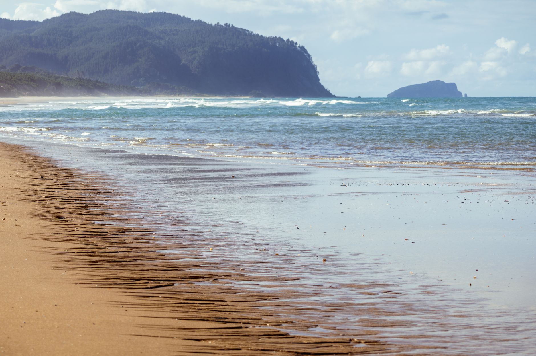 Peaceful sandy beach with waves and distant hills in Coromandel, New Zealand.