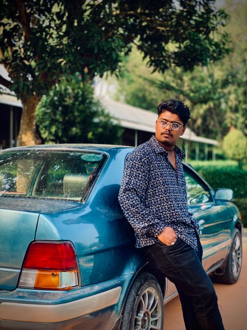 Free A stylish young man in glasses leaning on a blue car in an outdoor setting with greenery. Stock Photo