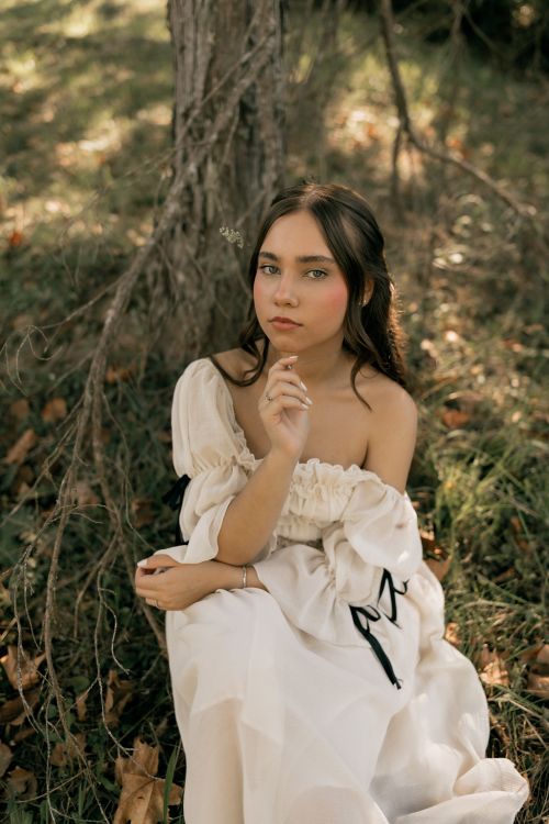 Free A serene portrait of a woman in a white dress, seated outdoors under a tree. Stock Photo