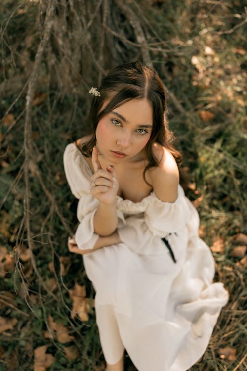 Free A contemplative young woman in a white dress sitting beneath a tree in a natural setting. Stock Photo