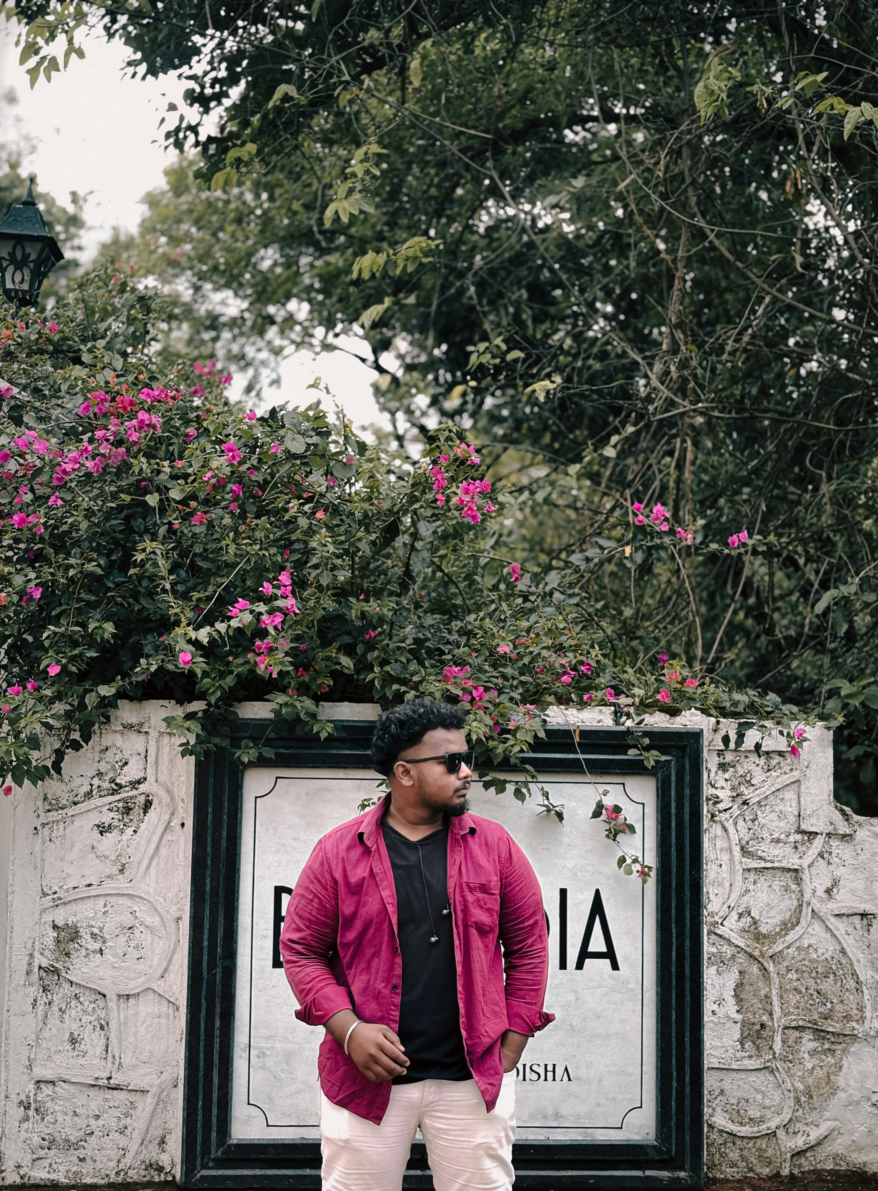 Man stands confidently in front of a floral bush with pink flowers, setting a stylish and relaxed outdoor vibe.