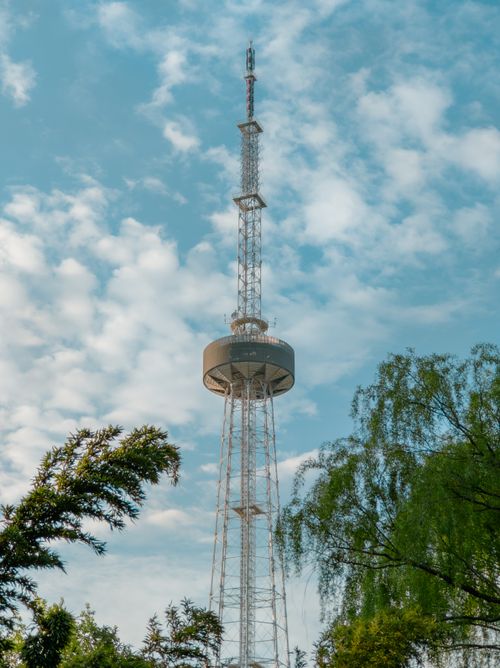 Free A tall communication tower reaching into a cloudy blue sky with green trees in the foreground. Stock Photo