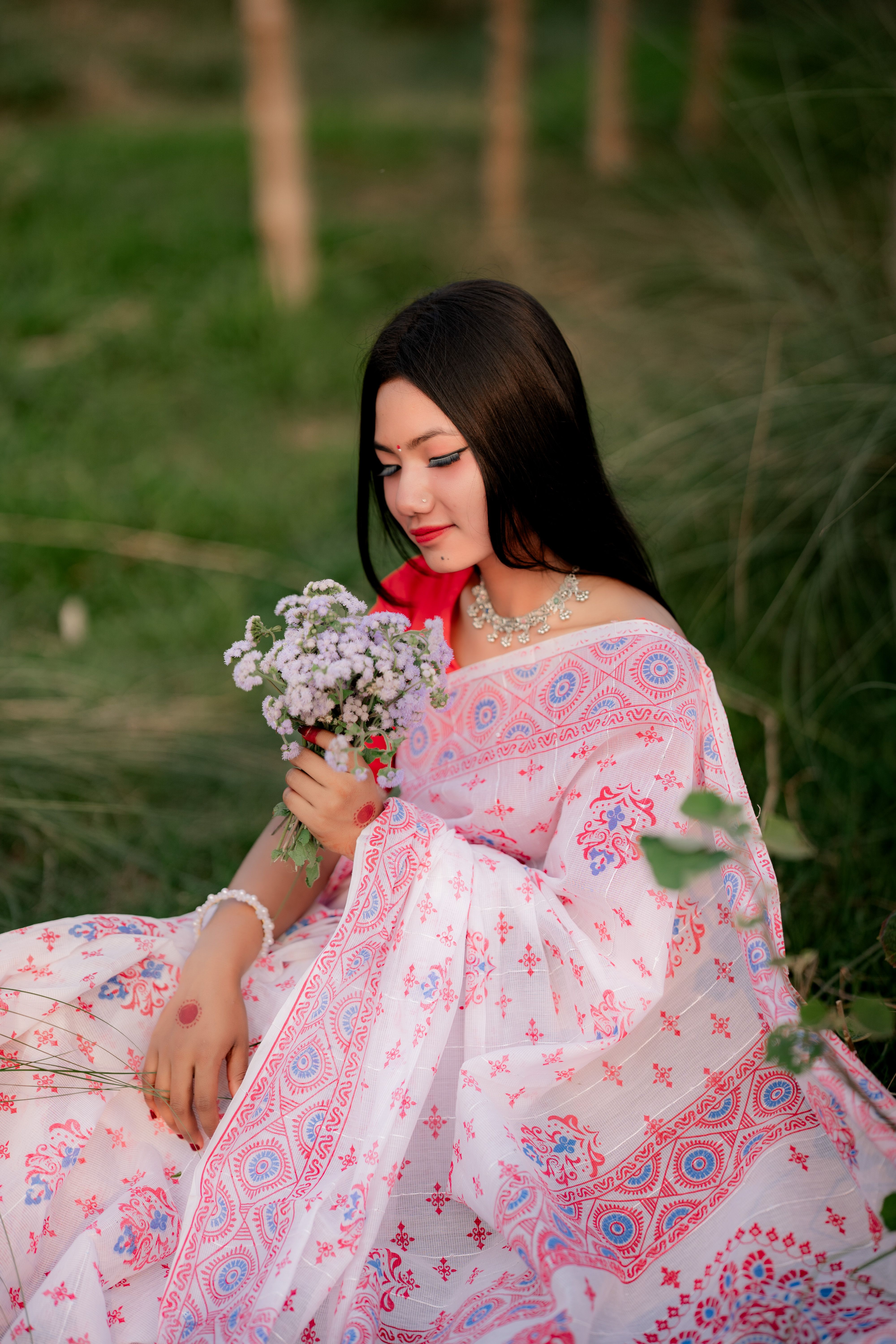 A woman in traditional Bangladeshi dress holding flowers, posing outdoors amidst nature.
