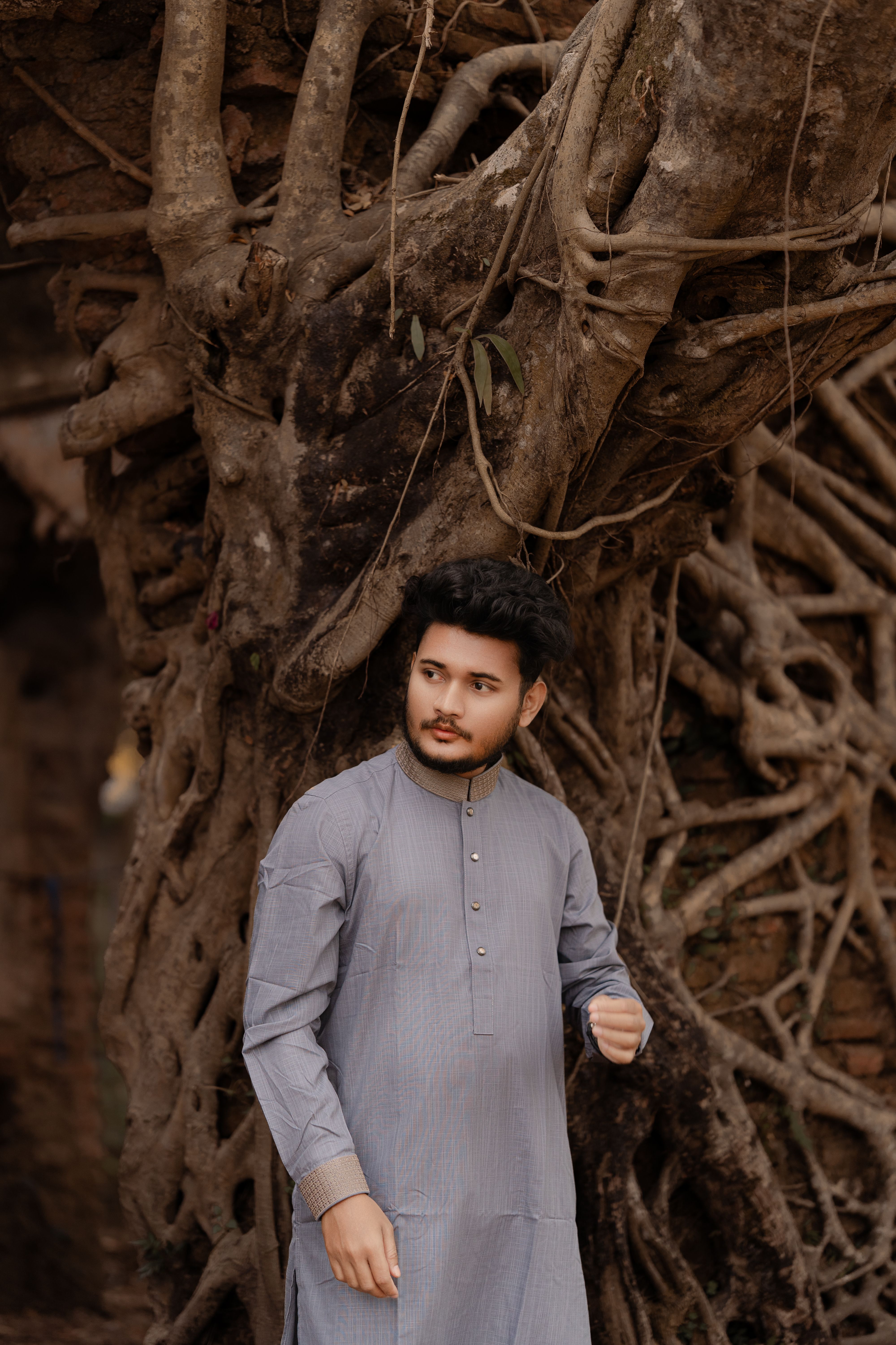 A young man wearing traditional Panjabi clothing stands confidently by a large tree in Bangladesh.