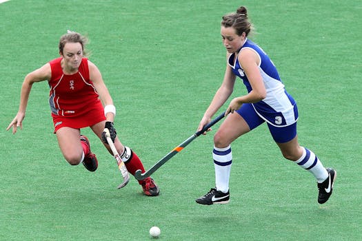 Two female athletes compete in a dynamic field hockey match on a green field.