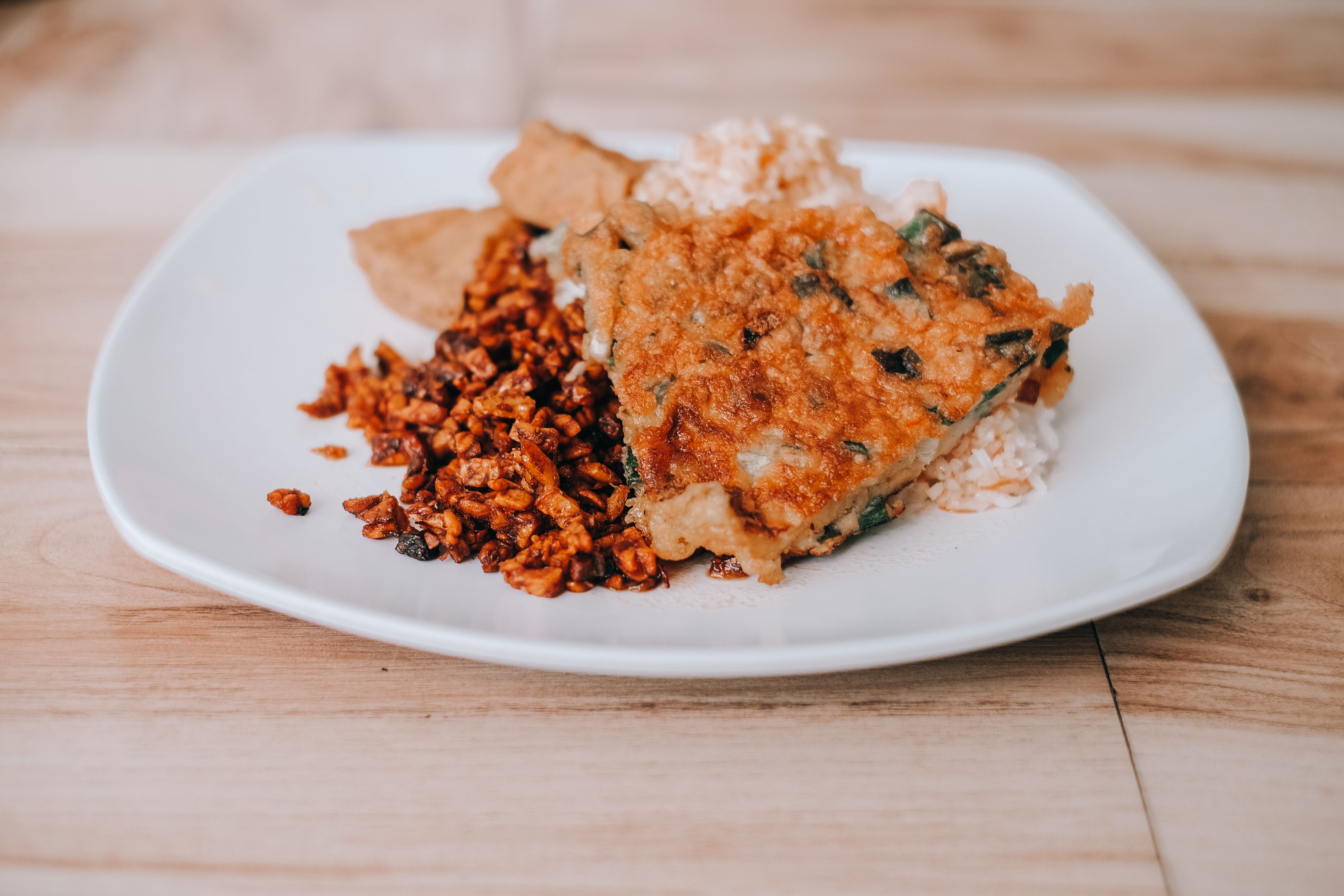 Delicious Indonesian tempeh dish with rice and crispy omelet on a white plate.