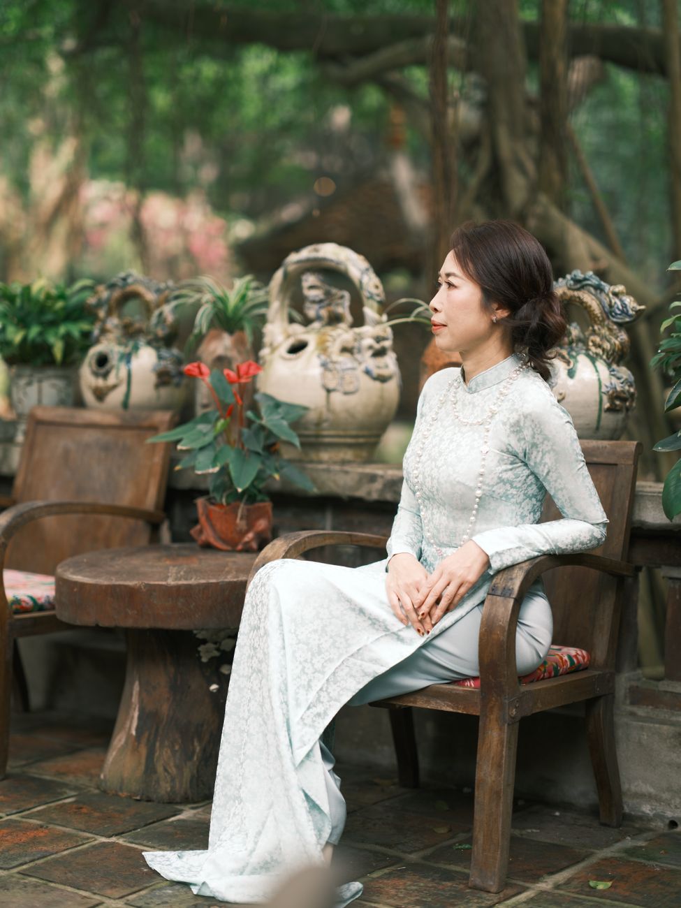 A woman in a traditional Vietnamese dress sits outdoors amidst greenery and pottery.