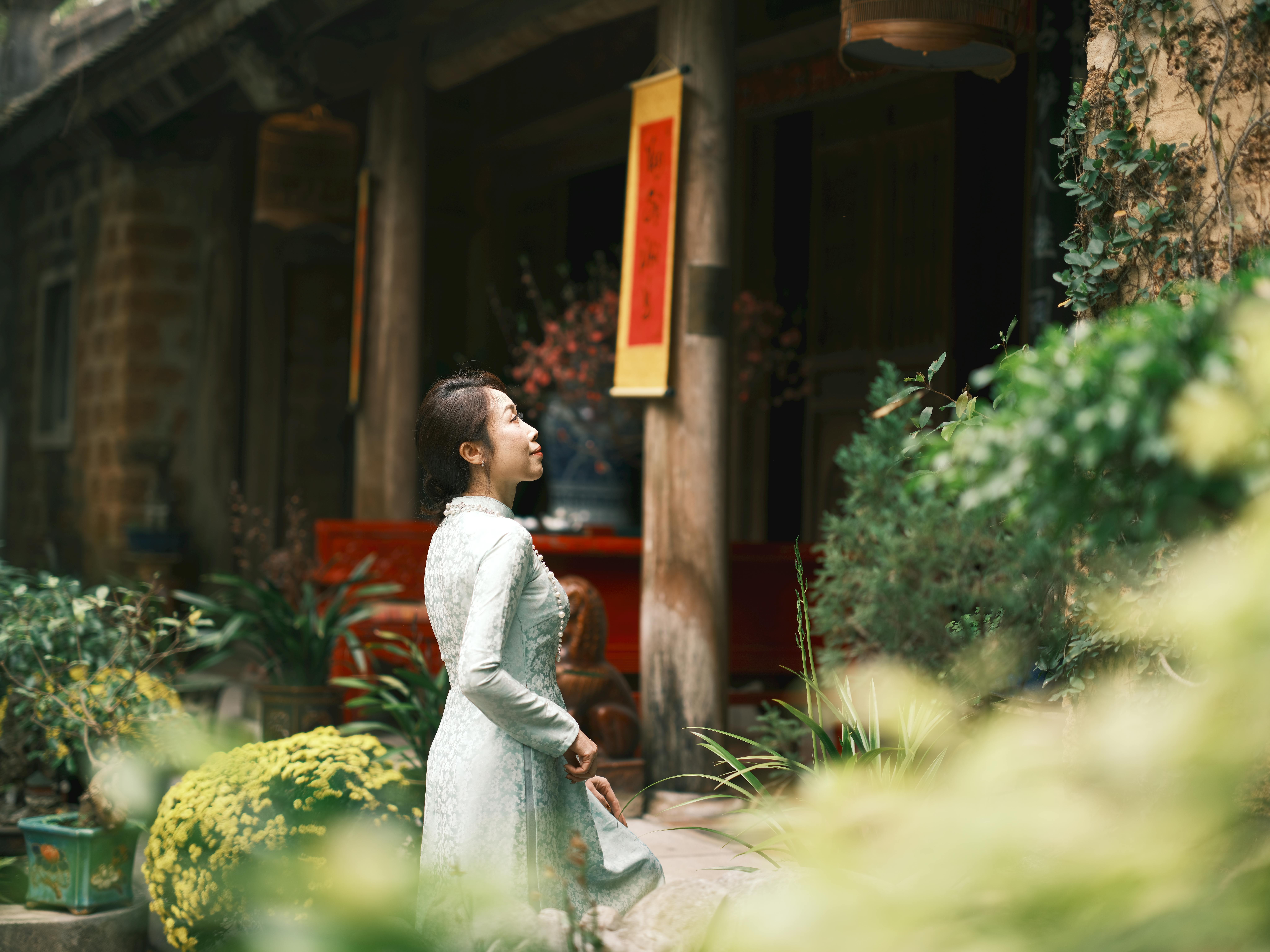 A woman in a traditional Vietnamese dress stands gracefully outside a rustic building surrounded by greenery.