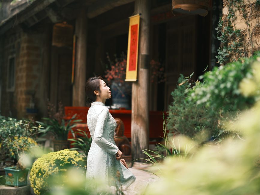 A woman in a traditional Vietnamese dress stands gracefully outside a rustic building surrounded by greenery.